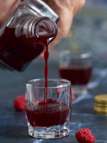 A hand holding a bottle of liqueur, pouring it into a small shot glass, another shot glass in the distance, bottle cap, and a couple of raspberries on a dark blue marble background.
