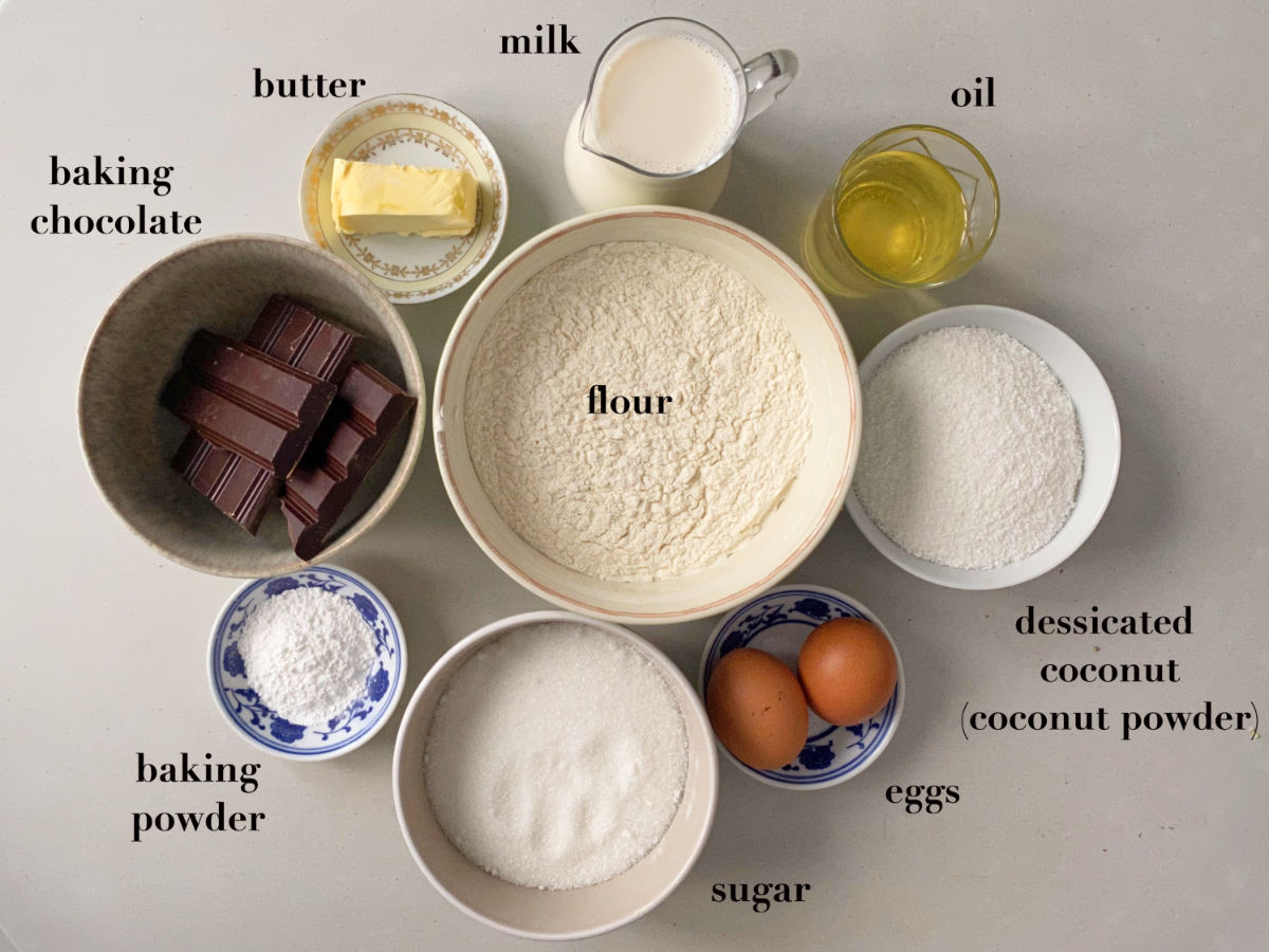 Ingredients on a gray background in white bowls: baking chocolate, butter, milk, oil, coconut, flour, baking powder, sugar and eggs. 