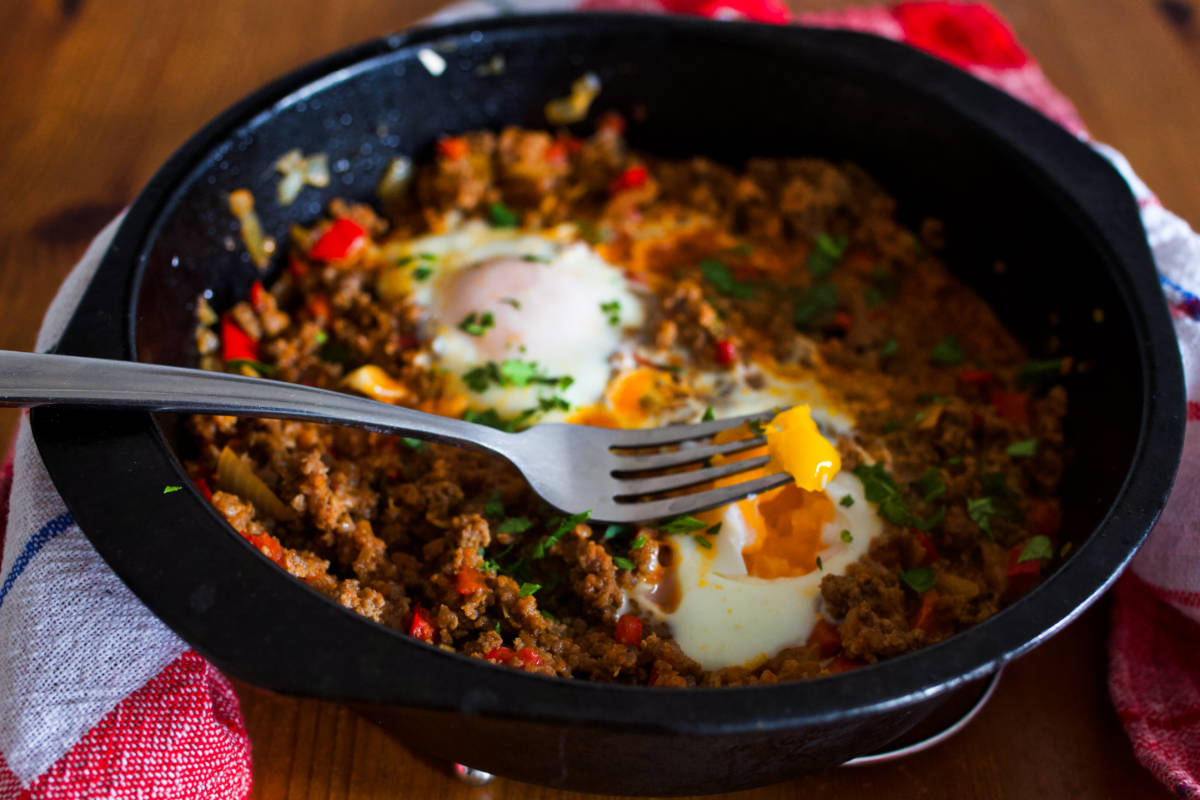 Black pan with two eggs and ground beef on a fork around which is a kitchen towel, all on a wooden table.