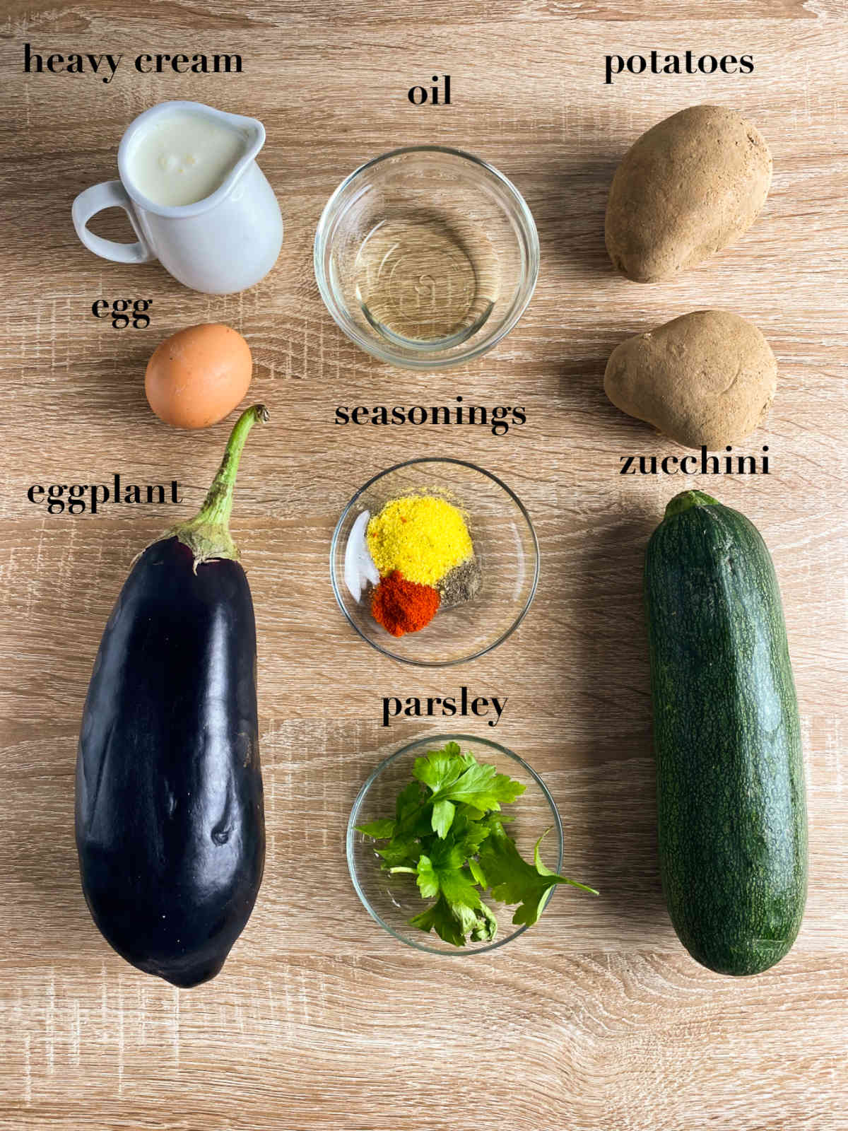 Ingredients for moussaka on a wood background (heavy cream, oil, potatoes, egg, seasonings, zucchini, eggplant and parsley).