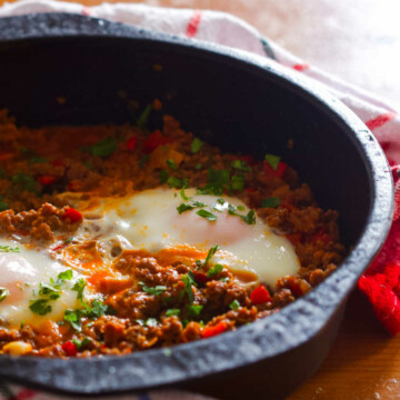 Black pan with two eggs and ground beef around which is a kitchen towel, all on a wooden table.