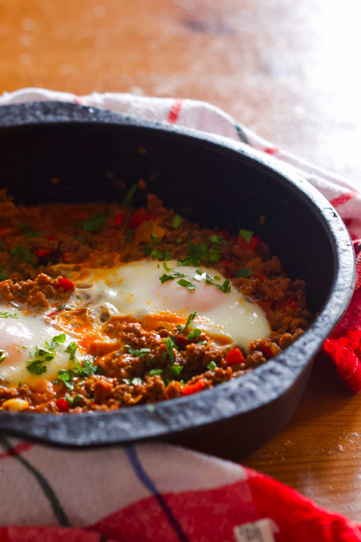 Black pan with two eggs and ground beef around which is a kitchen towel, all on a wooden table.