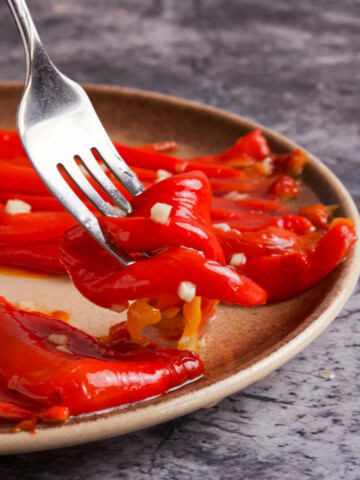 A fork holding red pepper salad above a plate on a marble table.