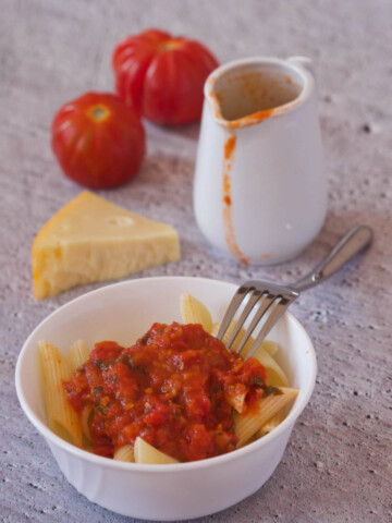 White bowl with pasta and tomato on a gray background together with two tomatoes cheese, fork and a pitcher with more tomato sauce.