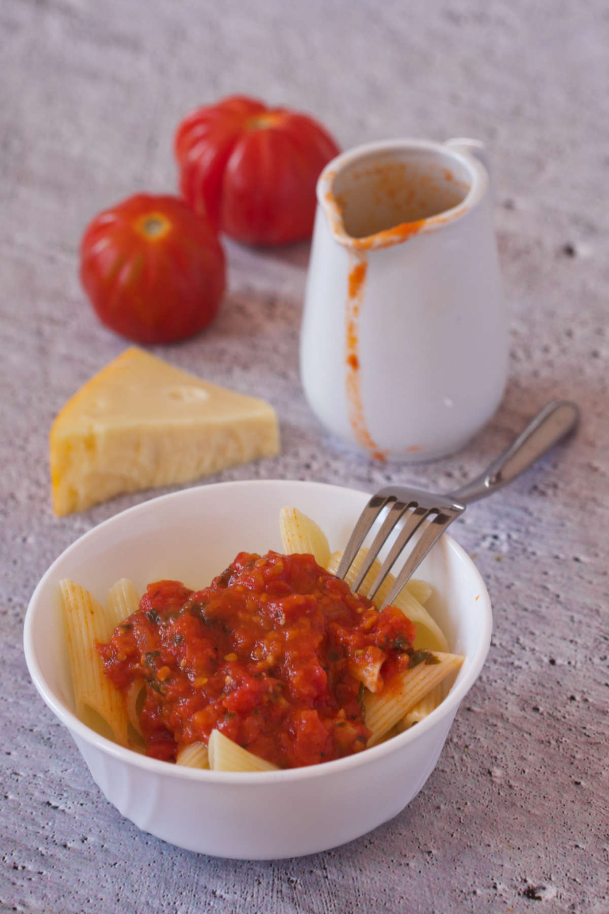 White bowl with pasta and tomato on a gray background together with two tomatoes cheese, fork and a pitcher with more tomato sauce.