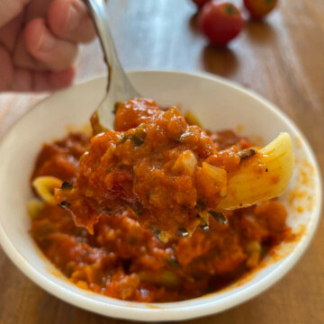 Hand holding fork with tomato sauce and pasta over white bowl.