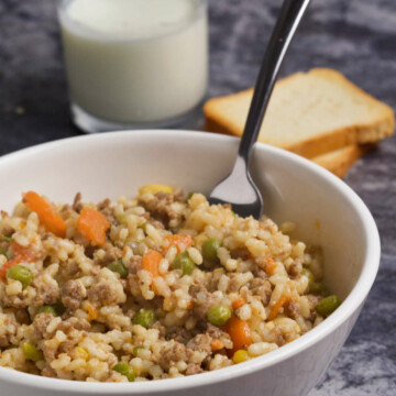 Bowl (white) with risotto and veggies with a fork in it, two pieces of toast and a glass with yogurt on gray marble background.