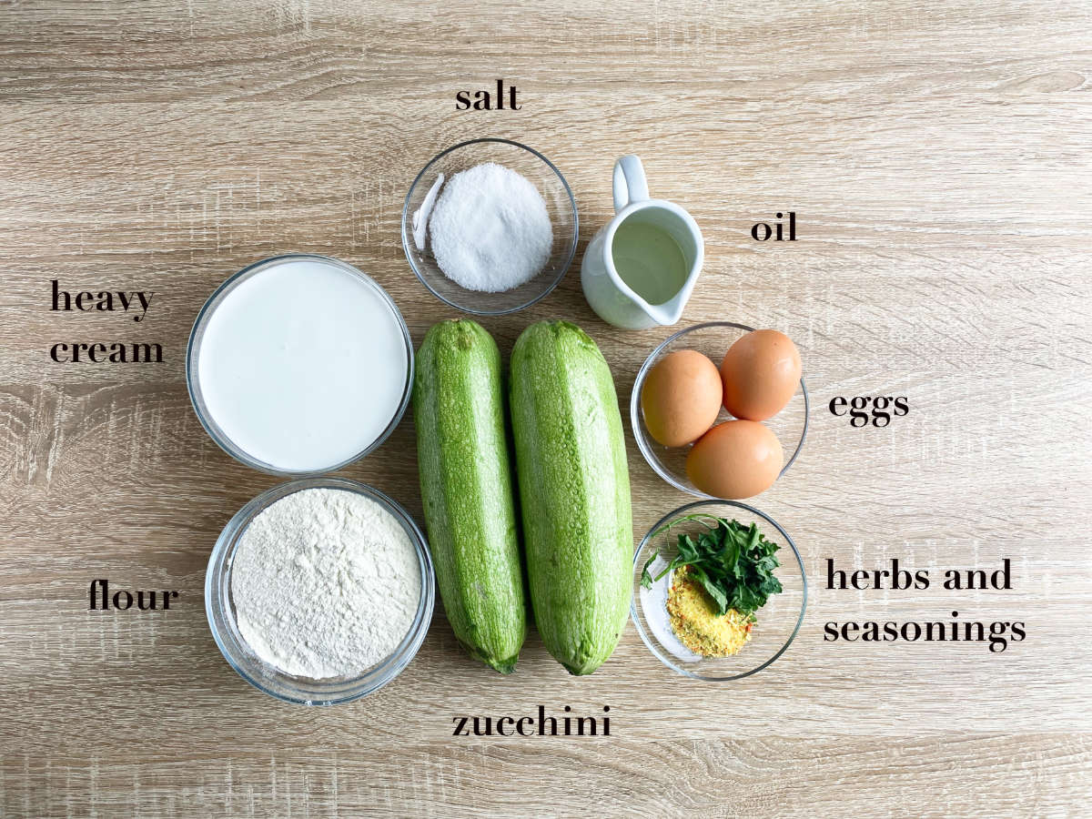 Ingredients on a wooden table: heavy cream, flour, salt, oil, eggs, herbs and seasonings in various glassware, and two zucchini. 