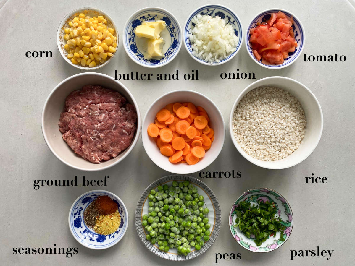Ingredients for the dish on a gray background: corn, butter, onion, carrot, tomato, ground beef, rice, seasonings, herbs, peas. 