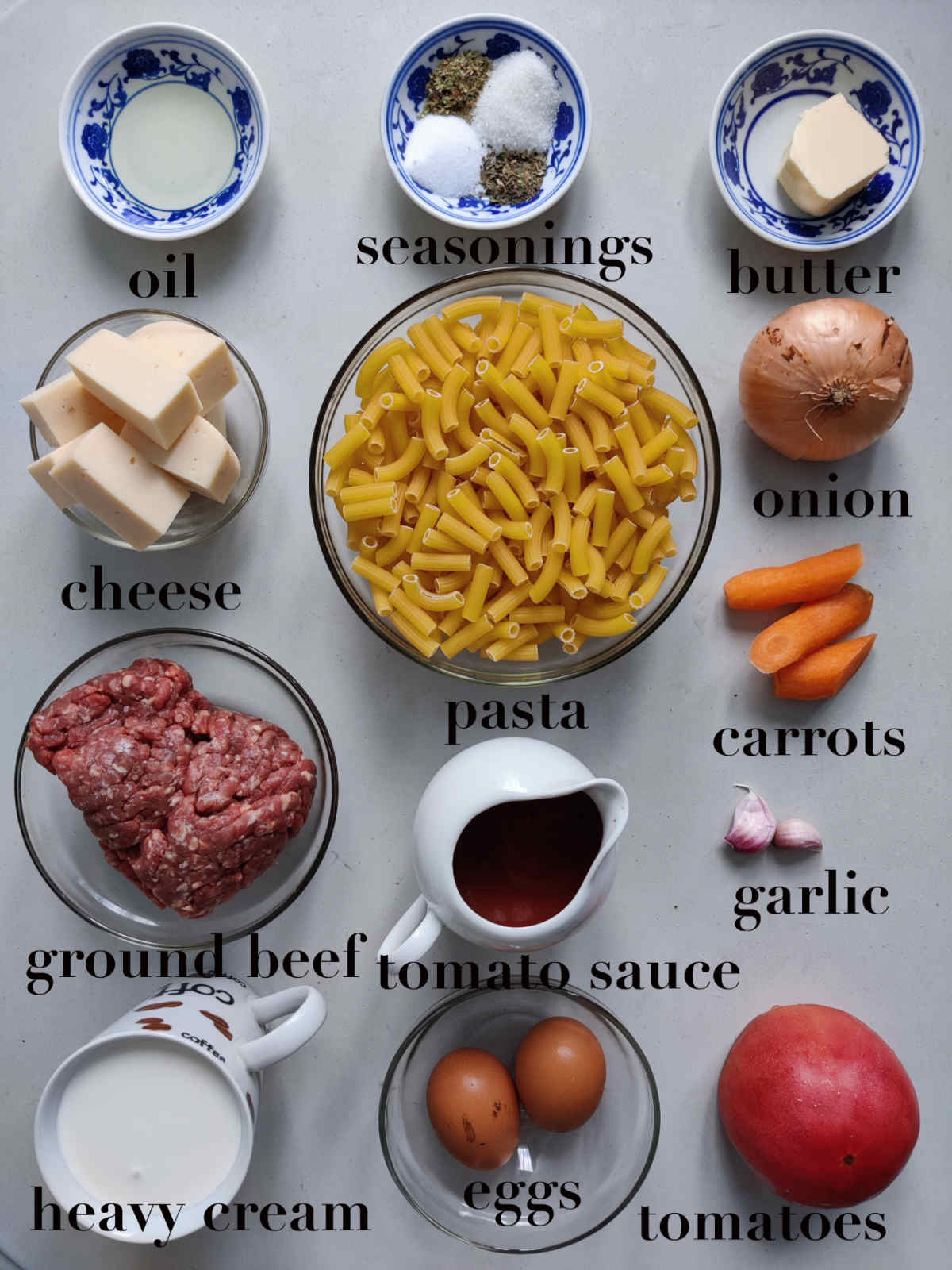 Overhead photo of ingredients for the recipe: oil, seasonings, butter, cheese, pasta, onion, carrots, ground  beef, tomato sauce, garlic, heavy cream, eggs and tomato.