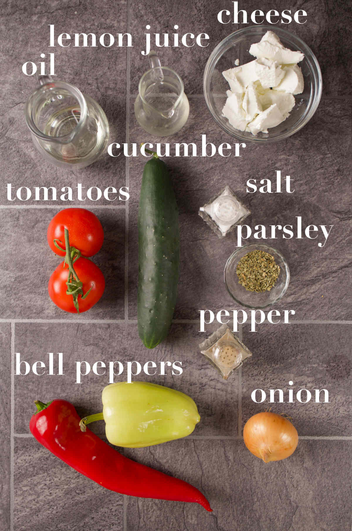 Oil, lemon juice, cheese, tomato, cucumber, peppers, onion and seasonings on a gray tabletop.