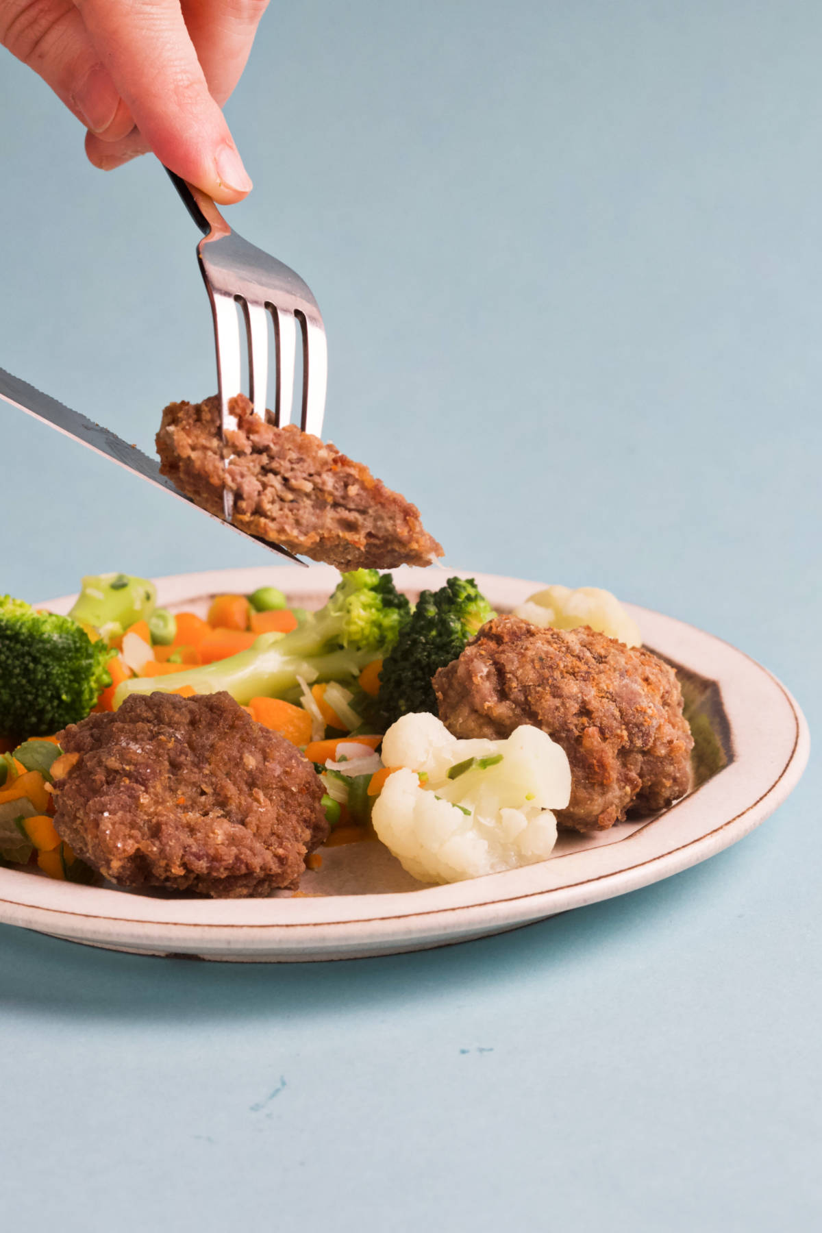 Fork and knife cutting into a rissole on a plate filled with veggies and rissoles.