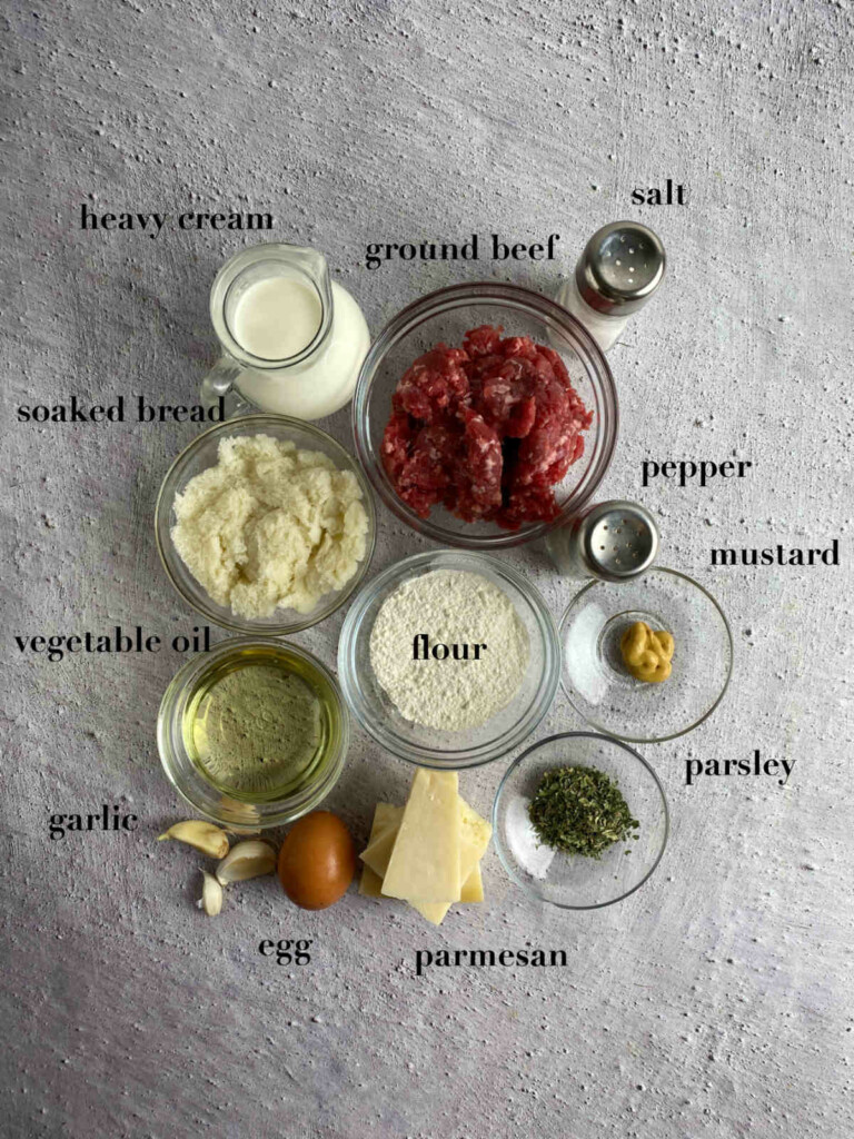 Ingredients for meatballs on gray background: heavy cream, bread, meat, seasonings, oil, egg, parmesan, flour.