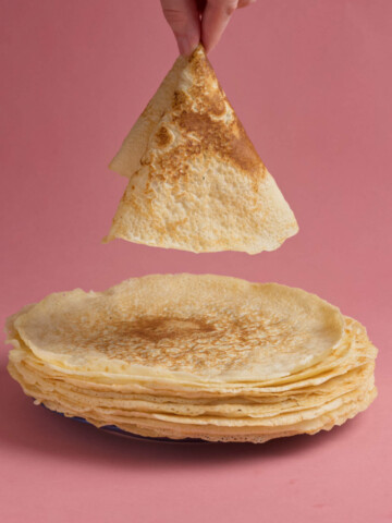 A hand holding a crepe above a plate of crepes on a pink background.