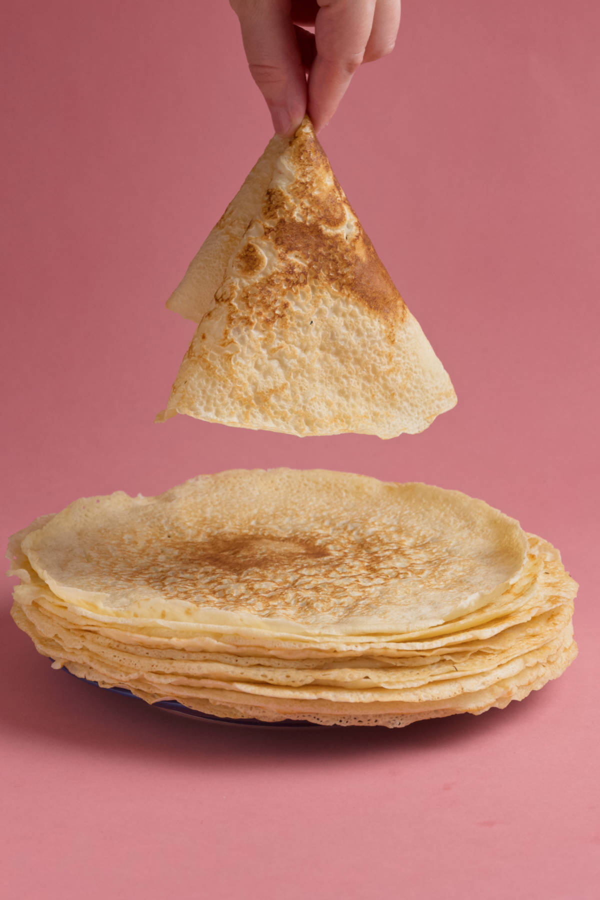 A hand holding a crepe above a plate of crepes on a pink background.