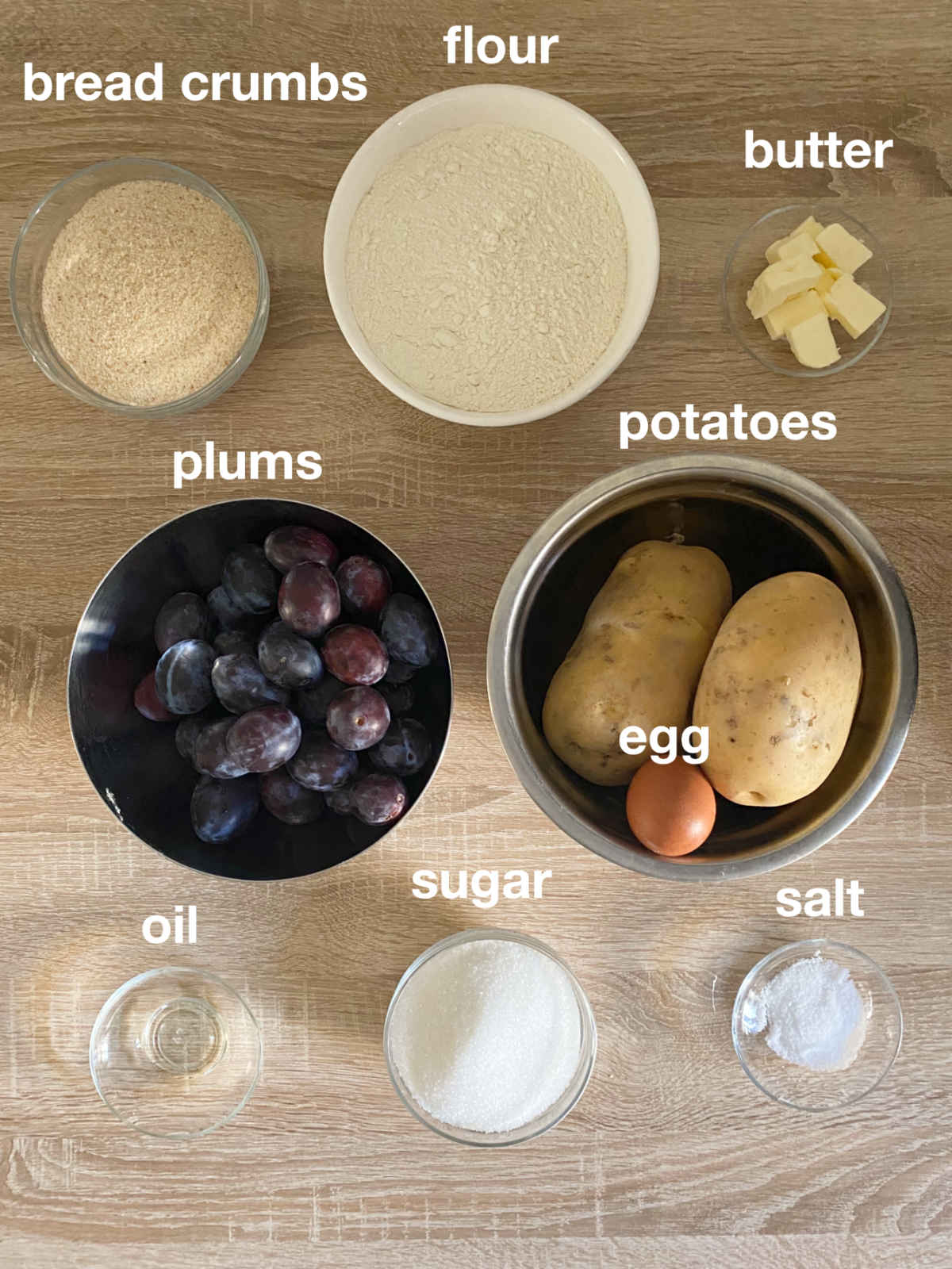 Overhead photo for ingredients for plum dumplings on a wooden table (bread crumbs, flour, butter, plums, potatoes, egg, oil, sugar, salt).