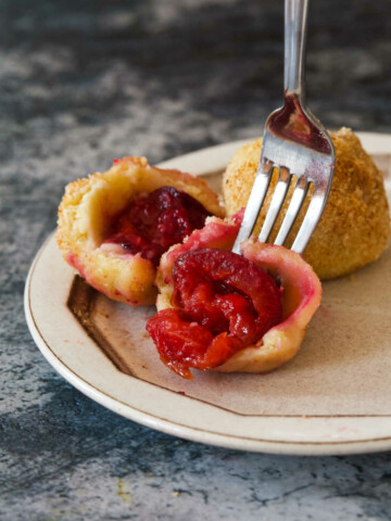 A fork holding half of a dumpling knedla on a white plate, with one and a half knedle in the background.