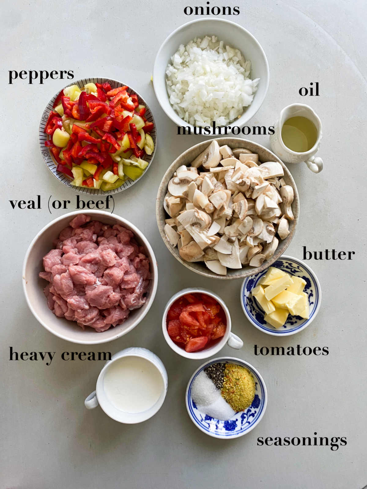 Nine ingredients in different bowls on a white background: peppers, tomatoes, onions, mushrooms, veal, oil, butter, heavy cream, tomato and seasonings.