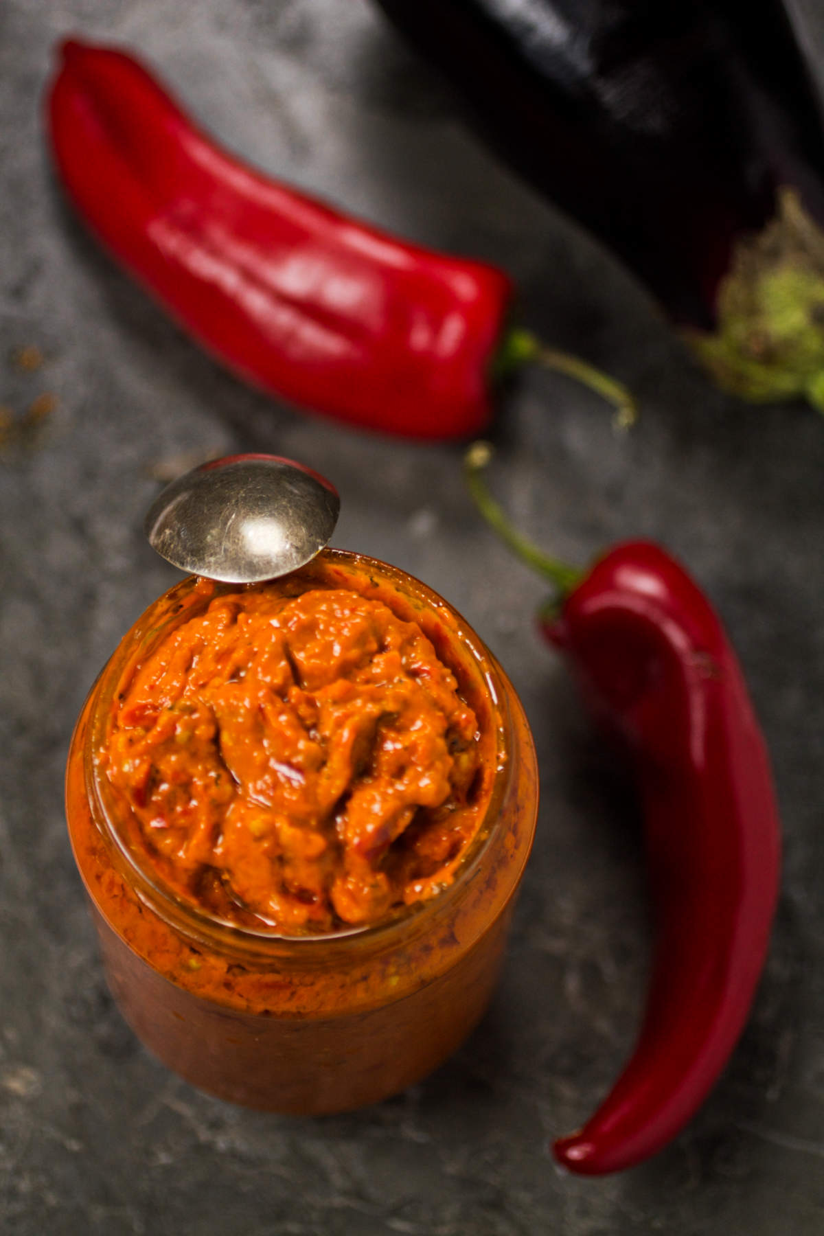 Ajvar in a jar on a gray background with eggplant and red peppers. 