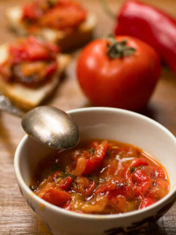 Sideways photo of pindjur in a bowl with a spoon and vegetables in the background.