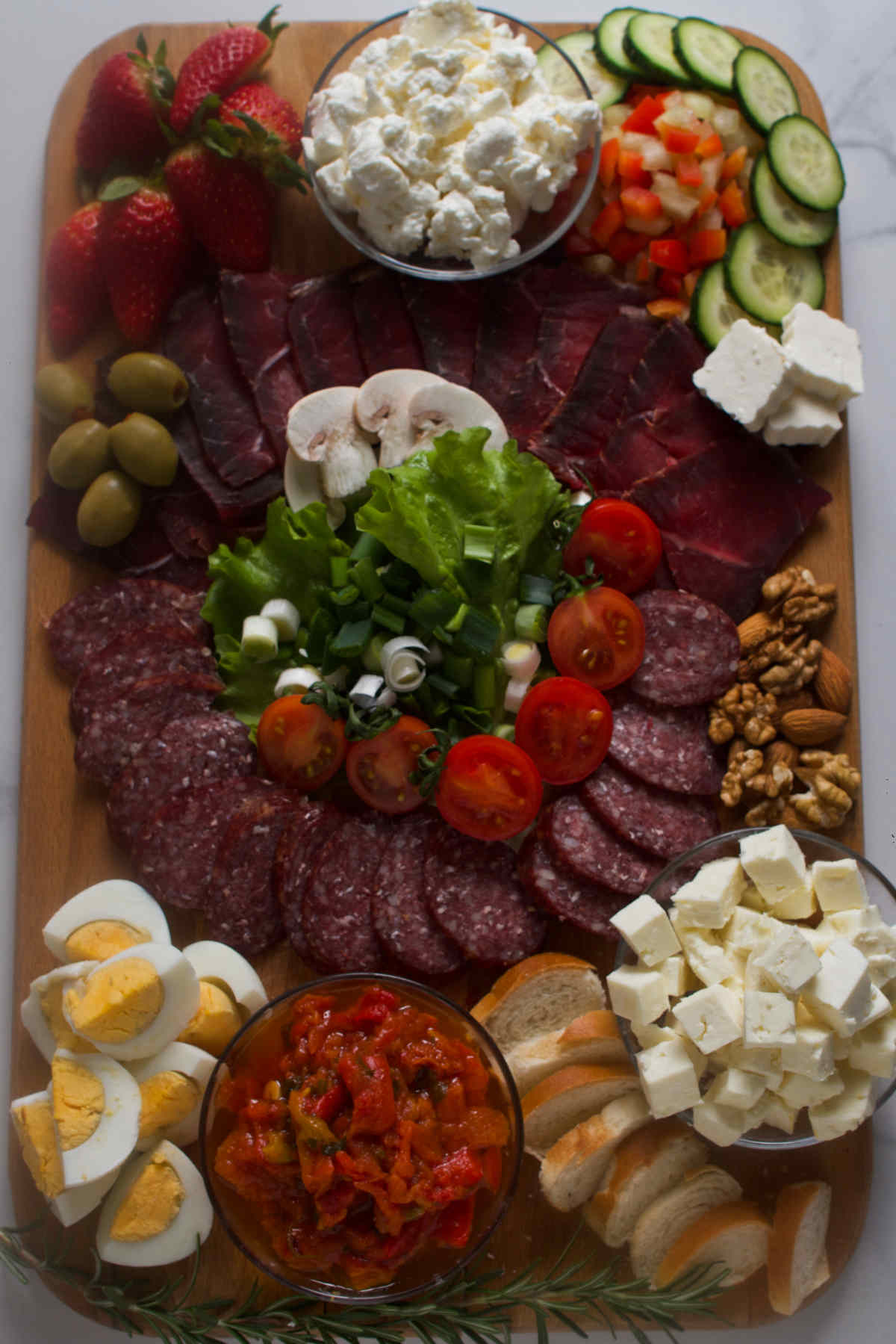 Rectangular wooden tray filled with different meats, cheeses, vegetables and nuts on a gray background. 
