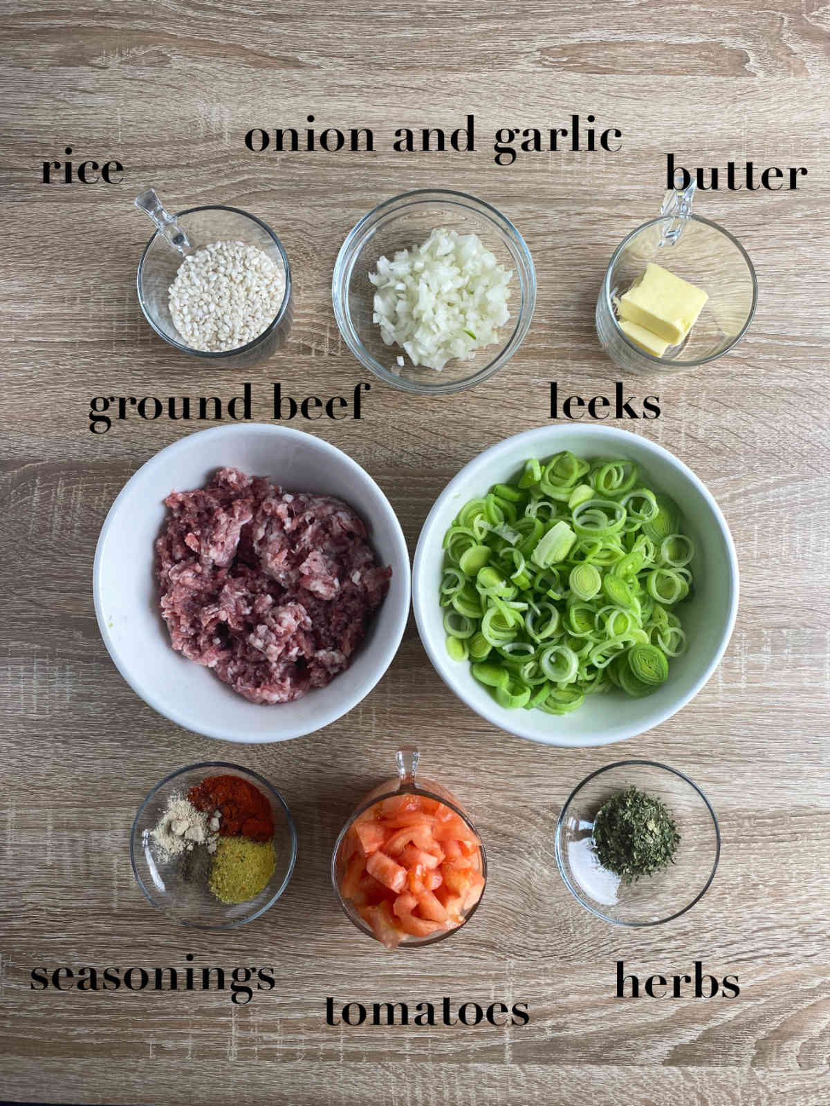 Eight bowls with ingredients for leek risotto on a wooden table: rice, garlic, onion, butter, tomato, seasonings, herbs, ground beef and leeks.