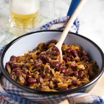 A blue pan filled with onions and kidney beans, and a jug of beer on a marble background; wooden spoon, kitchen towel and bread in the background.