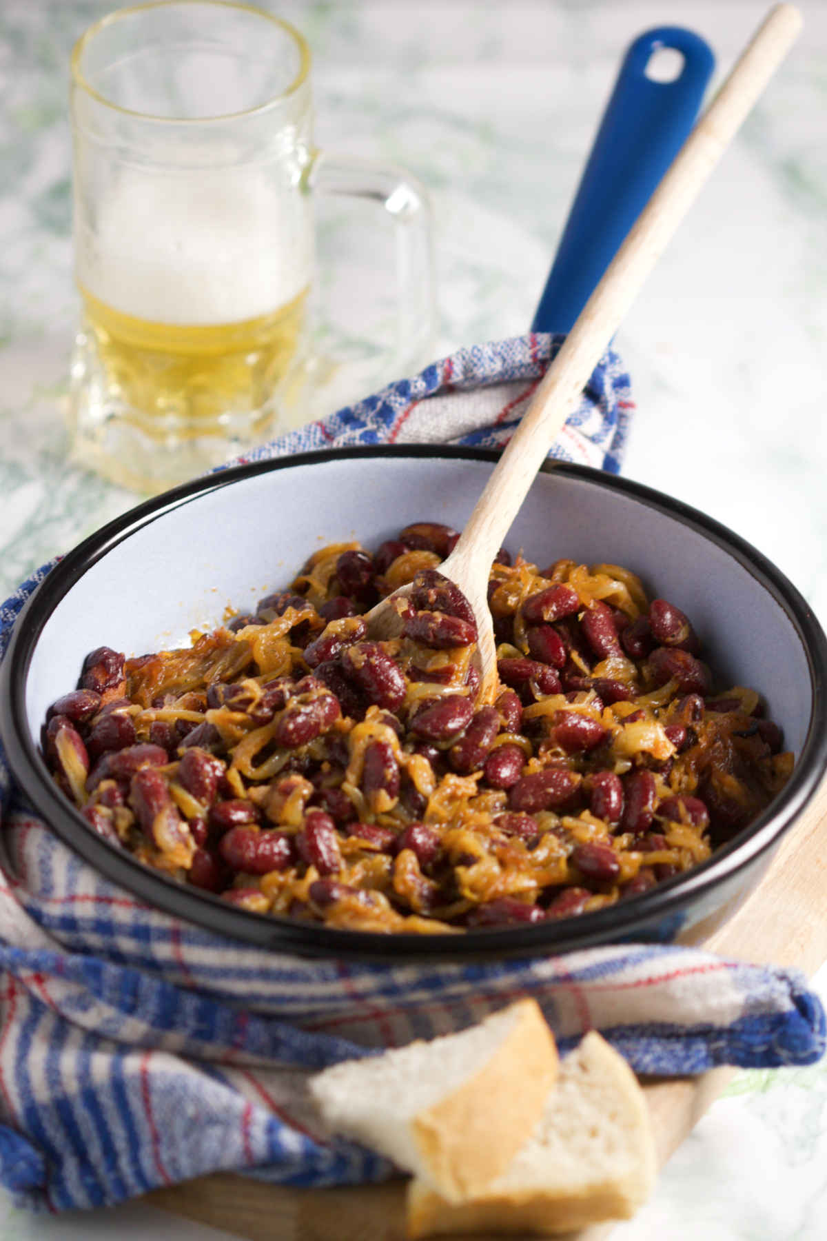 A blue pan filled with onions and kidney beans, and a jug of beer on a marble background; wooden spoon, kitchen towel and bread in the background. 