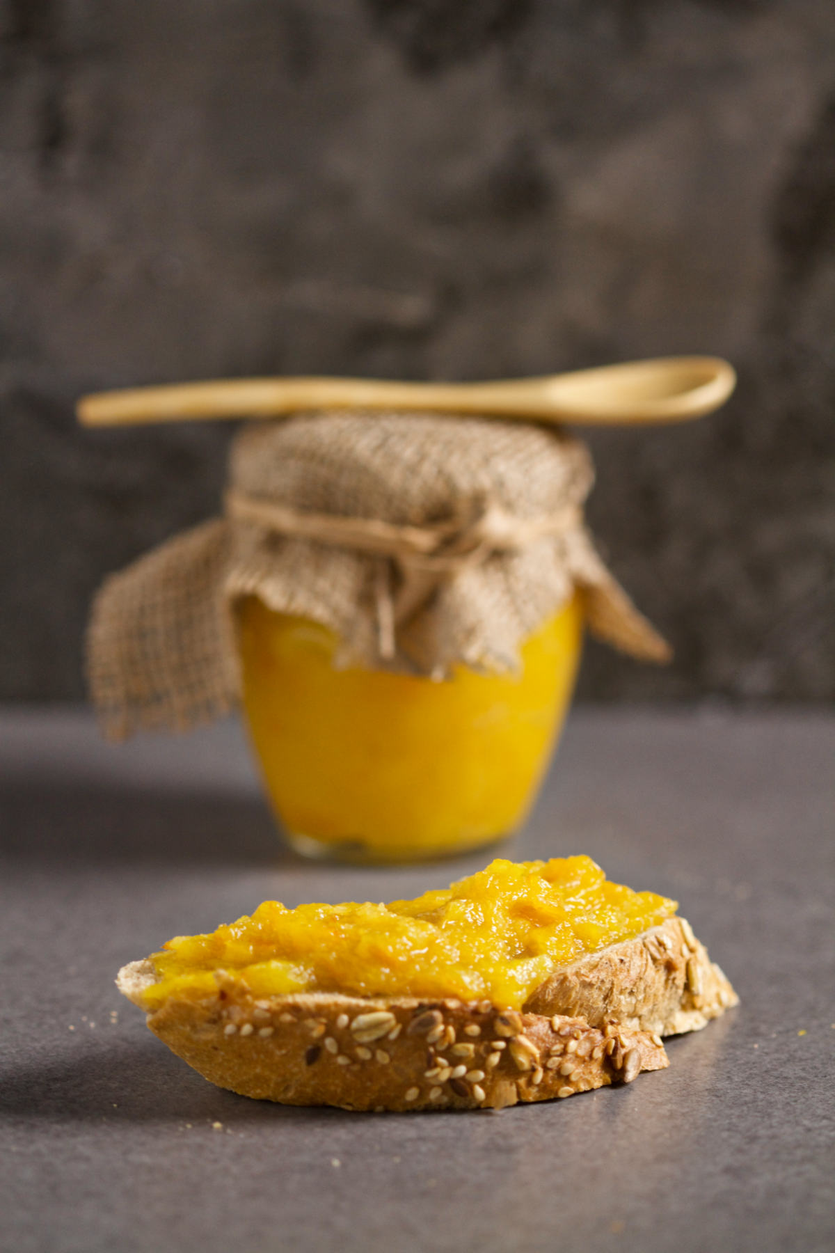 A piece of integral bread with marmalade and a jar of marmalade with a wooden spoon on top, on a gray background.