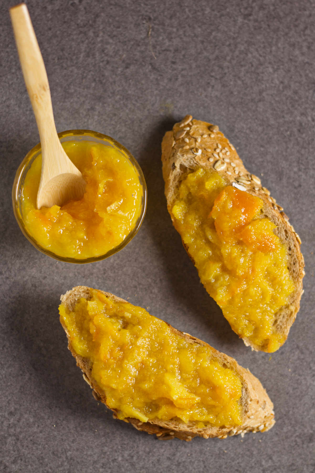 Two slices of integral bread with orange marmalade, and a bowl with orange marmalade and a wooden spoon on a gray background.