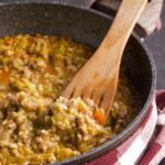 Black pan with risotto and wooden spoon, plaid kitchen towel, all on a black background.