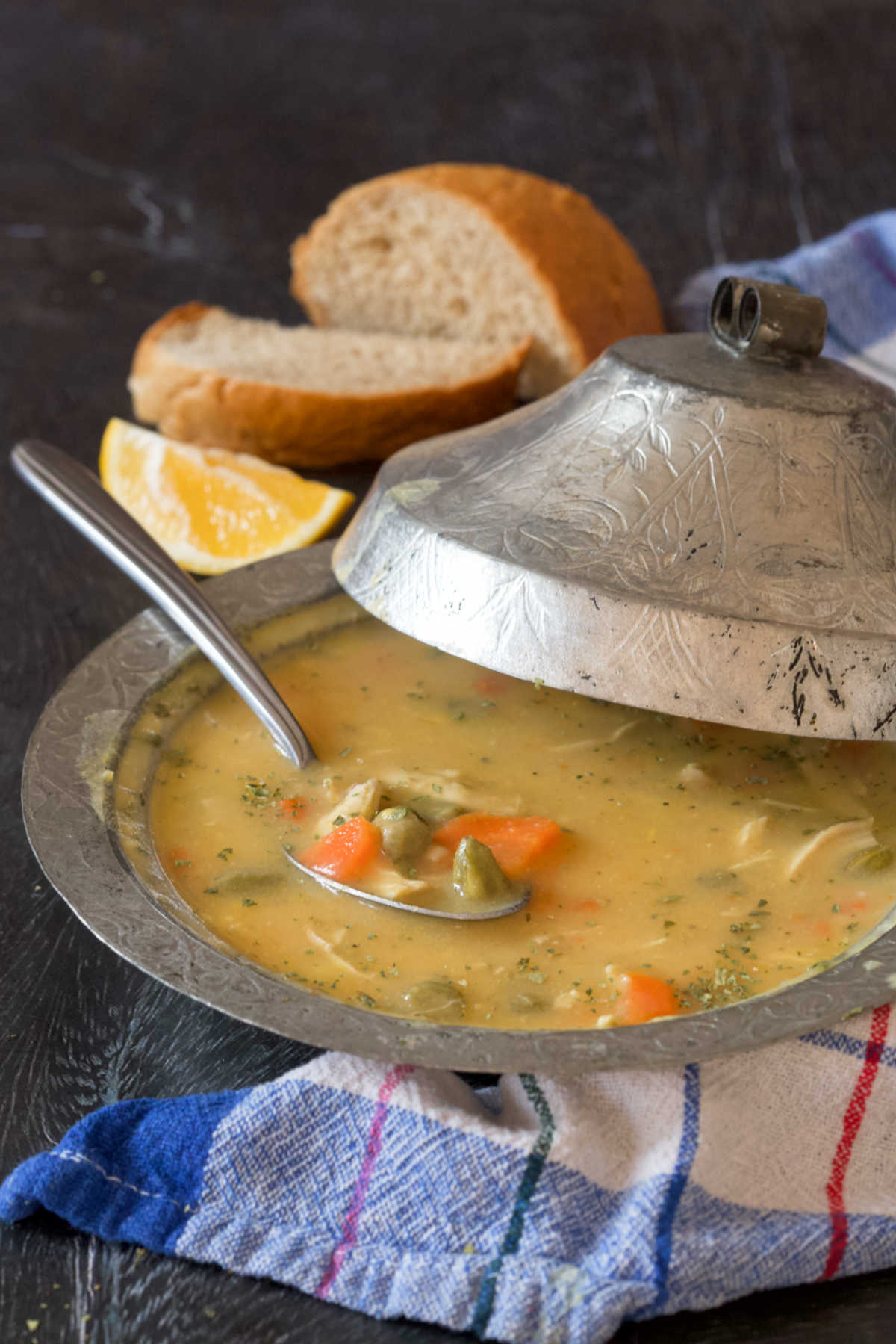 Soup in a silver bowl with a lid, a kitchen cloth, two slices of bread, a lemon wedge and a spoon.