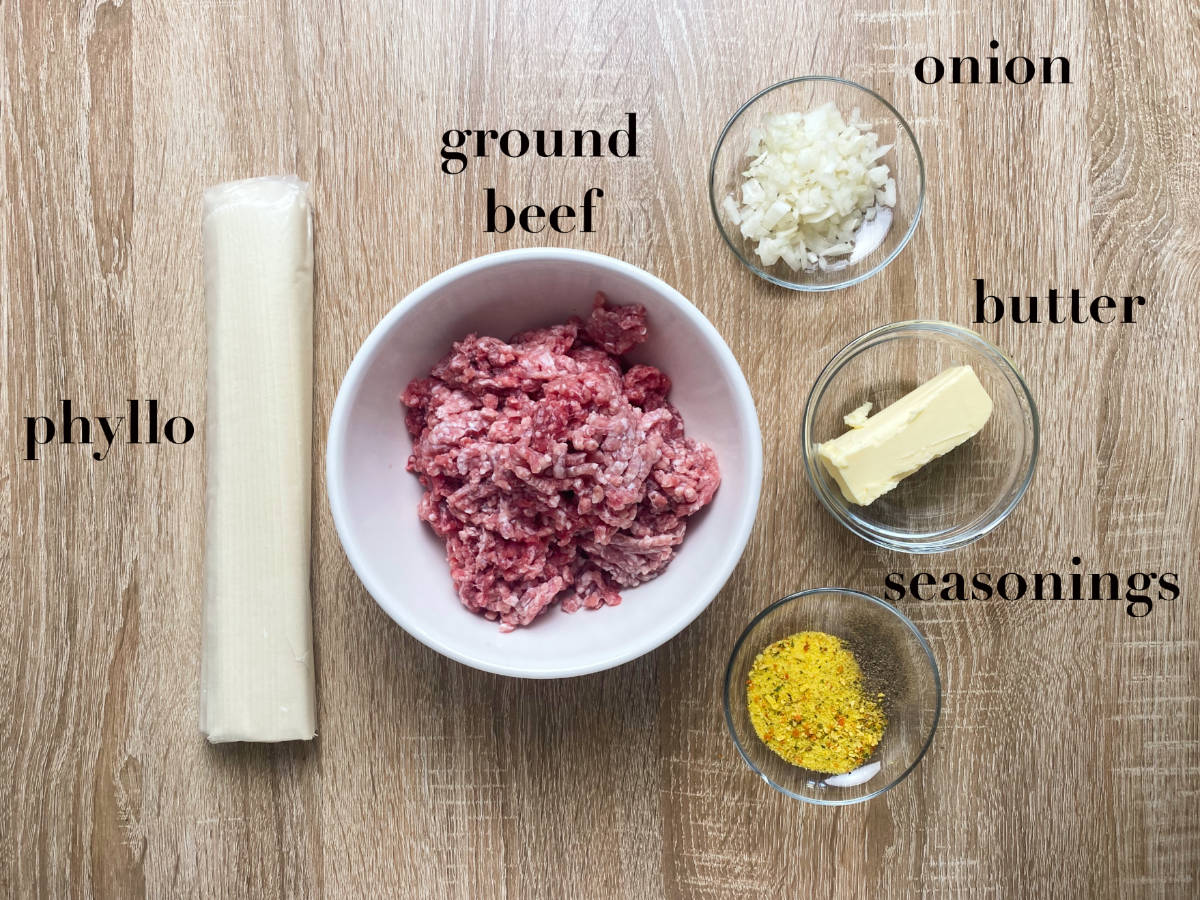 Ingredients on wooden table: phyllo, ground beef, onion, butter, seasonings. 
