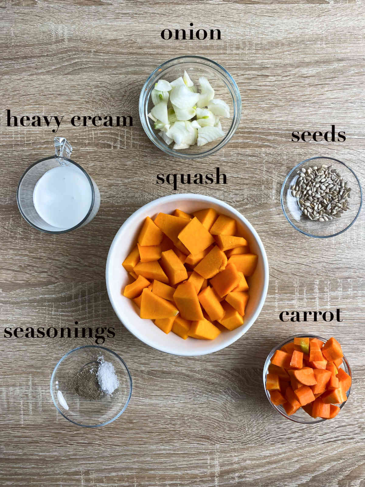 Six bowls with different ingredients on a table: heavy cream, onion, seeds, squash, carrot and seasonings. 