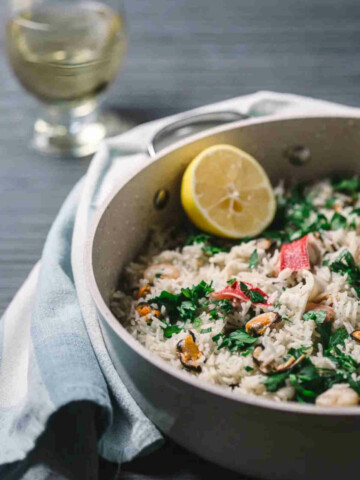 Pan with seafood risotto and slice of lemon on a gray background, together with a glass of wine and kitchen towel.