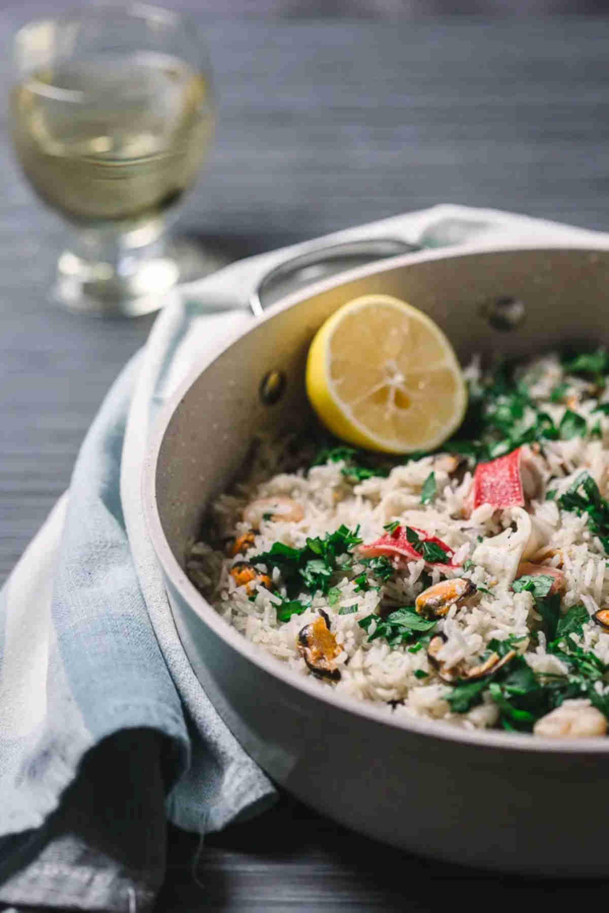 Pan with seafood risotto and slice of lemon on a gray background, together with a glass of wine and kitchen towel. 