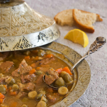 Silver bowl with a lid filled with a spoonful of the sauce, a lemon wedge, and a slice of bread on gray background.