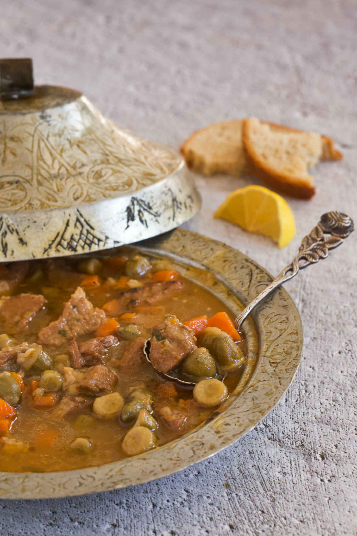 Silver bowl with a lid filled with a spoonful of the sauce, a lemon wedge, and a slice of bread on gray background.