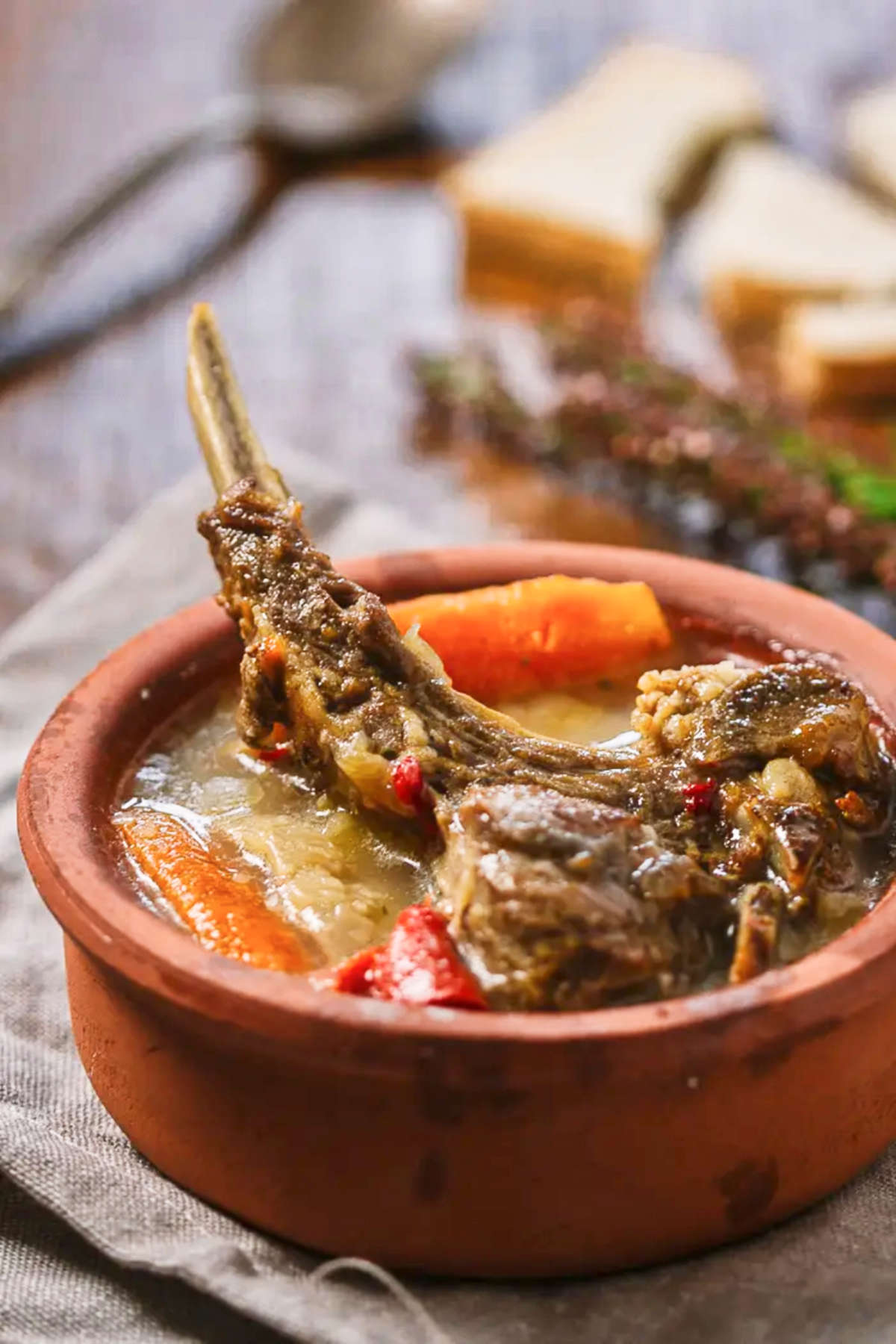 A clay pot with a lamb chop, and carrot stew, a spoon, and bread on a gray purple background. 