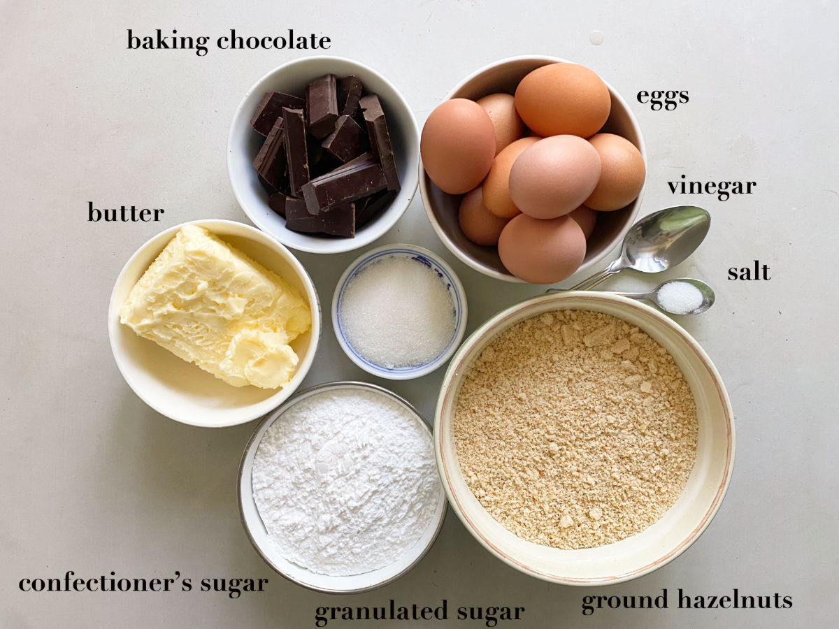 Eight ingredients in spoons and bowls on a white background: chocolate, eggs, butter, sugar, vinegar, salt and hazelnuts.