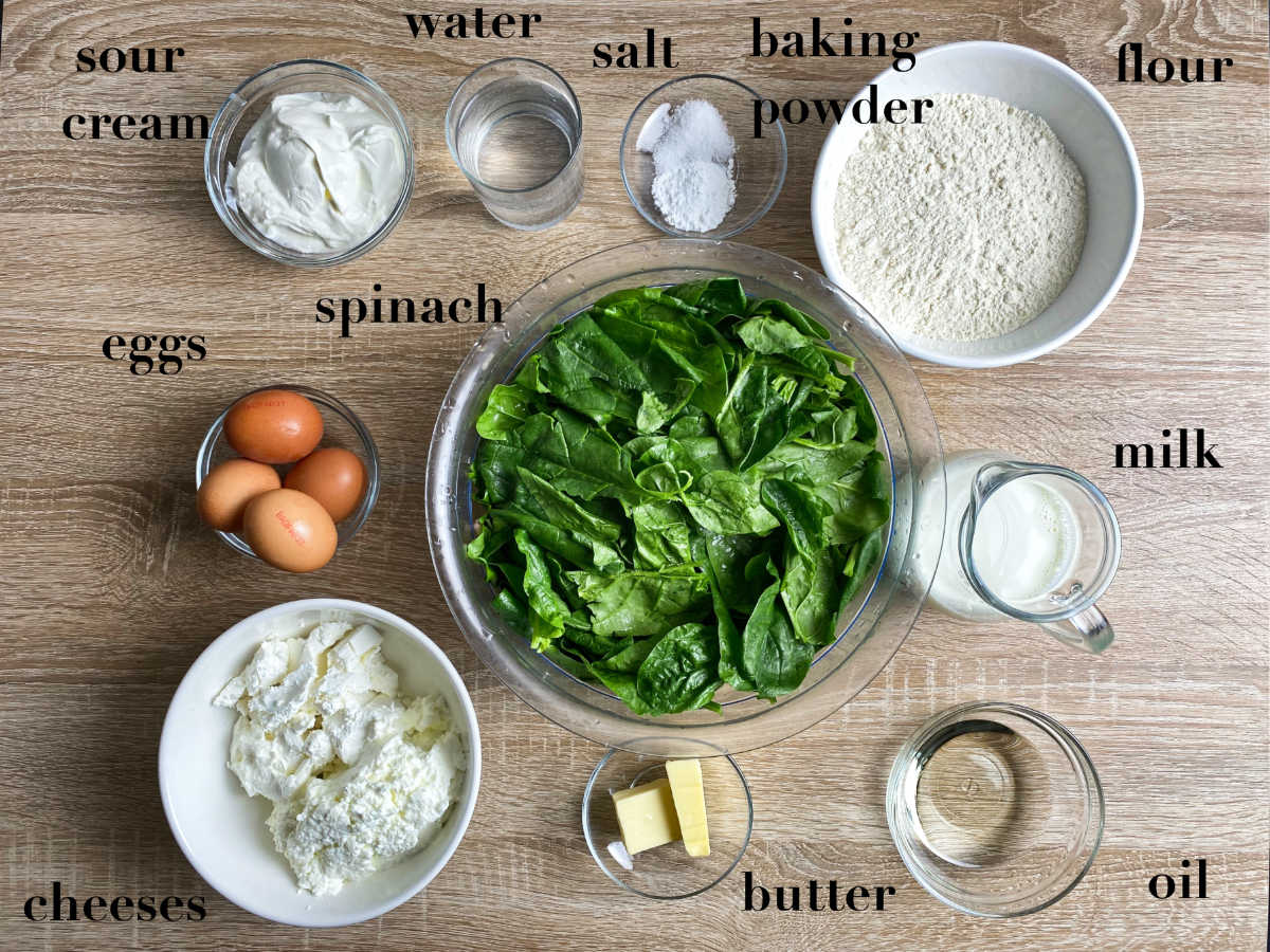 Dish ingredients on a wooden table: salt, sour cream, baking powder, eggs, cheeses, spinach, oil, butter, milk, flour and water. 