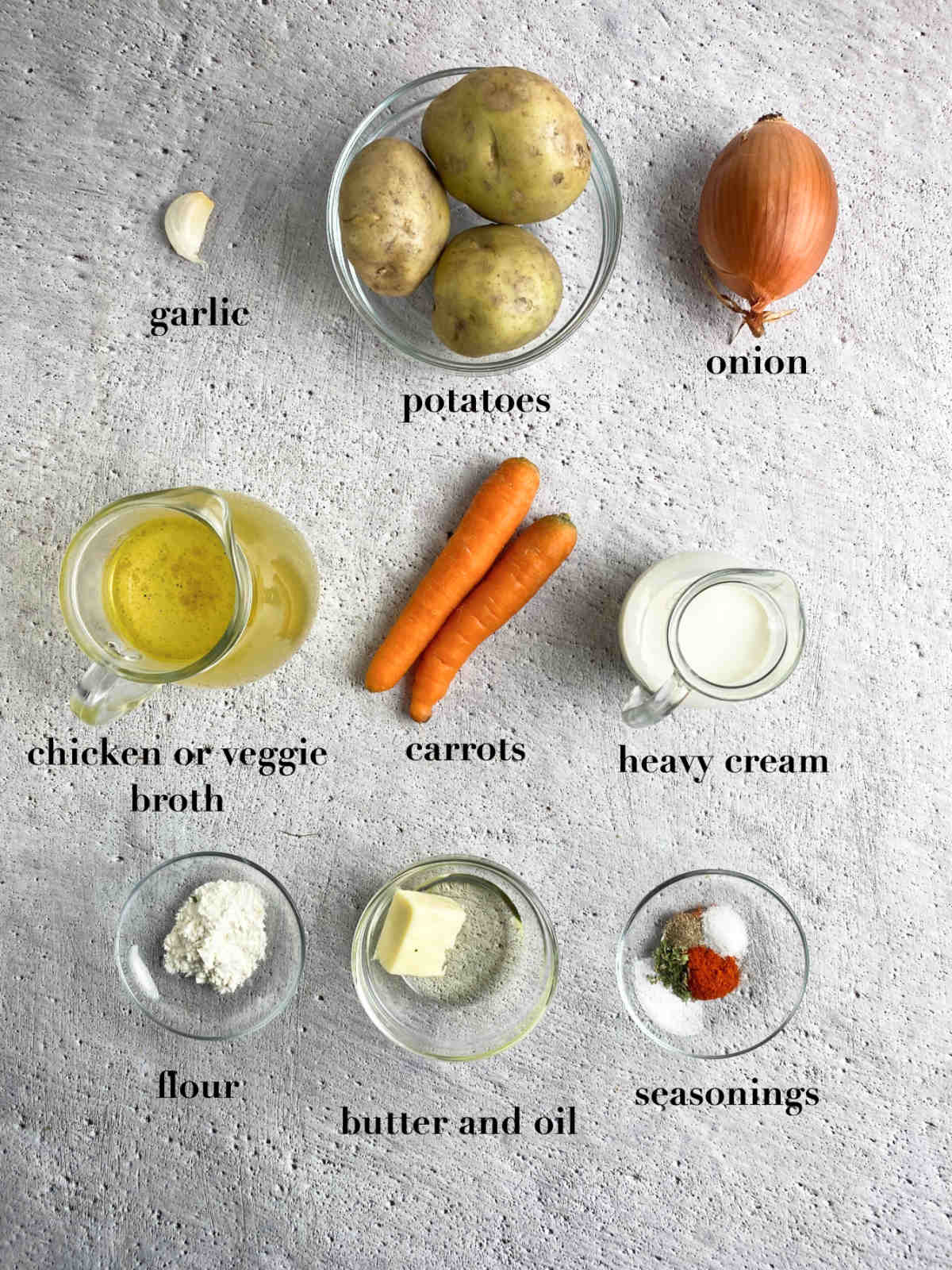 Ingredients for the soup on a gray table: garlic, onions, potatoes, broth, carrots, heavy cream, flour, butter, oil and seasonings. 