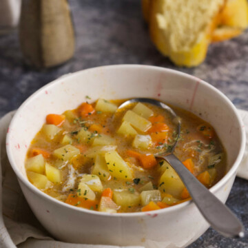 A bowl with potato and carrot soup, a spoon, bread, salt and pepper on a gray background.