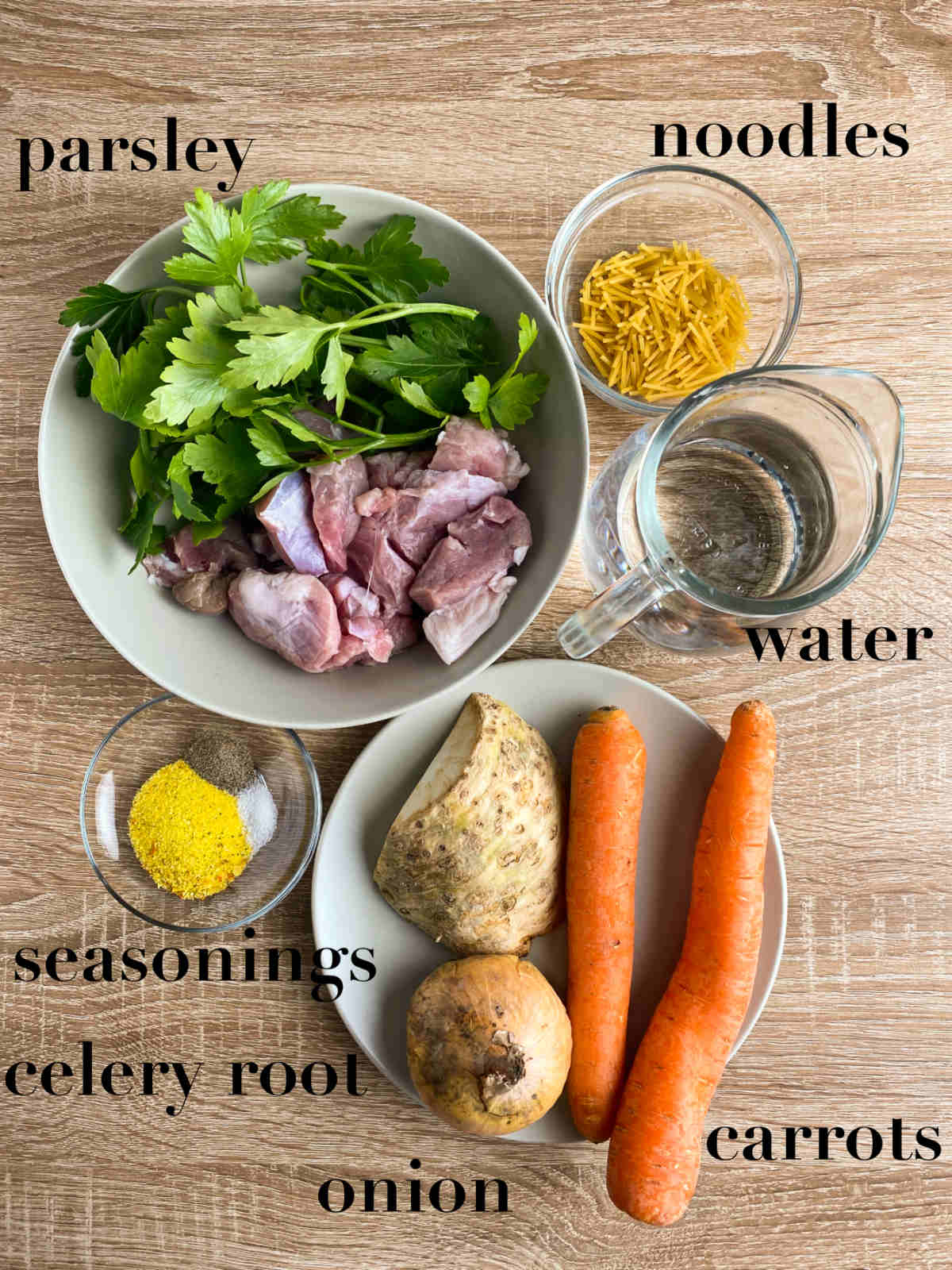 Ingredients for veggie beef soup on a table (carrot, parsley, beef, noodles, water, seasonings, onion, celery). 