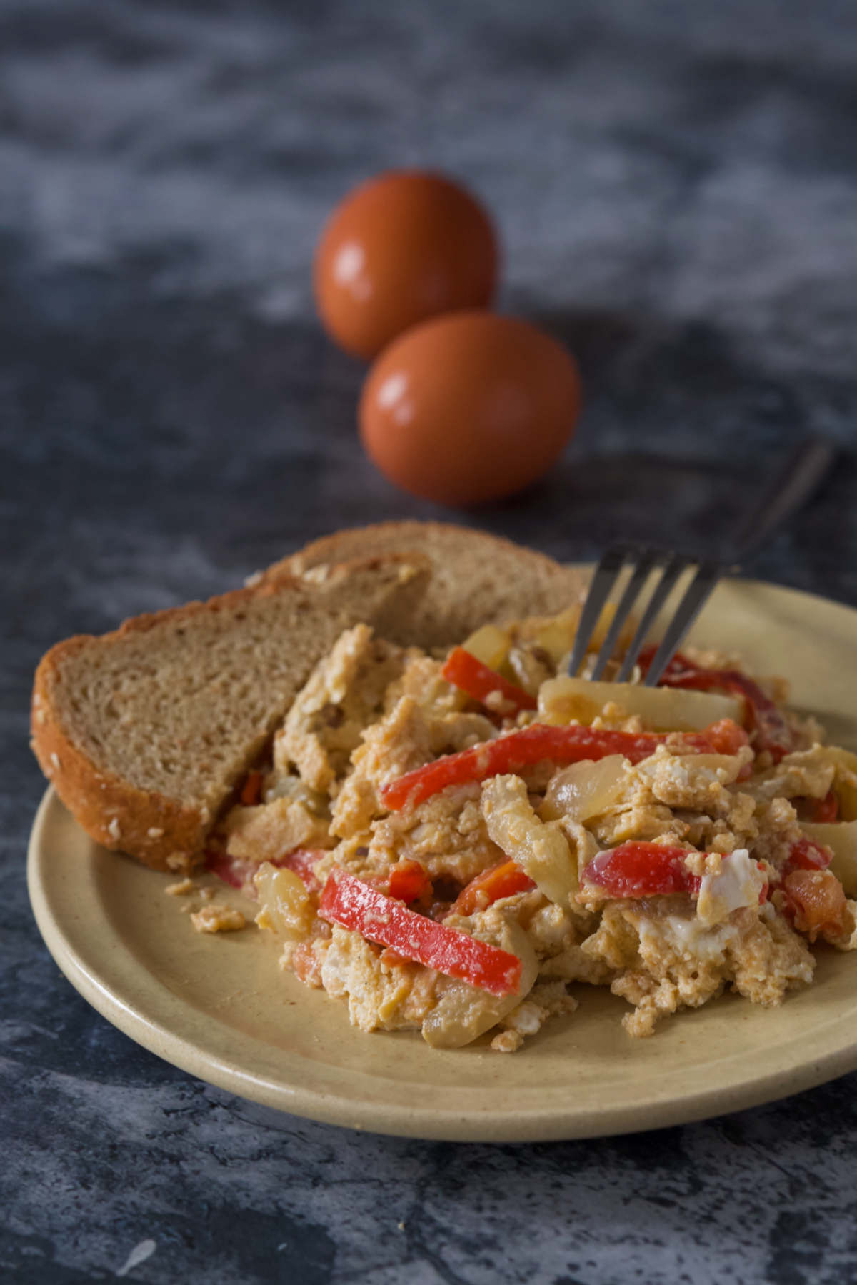 A plate with eggs and peppers (satarash) with two halves of integral bread with a fork and two eggs on a dark marble background.