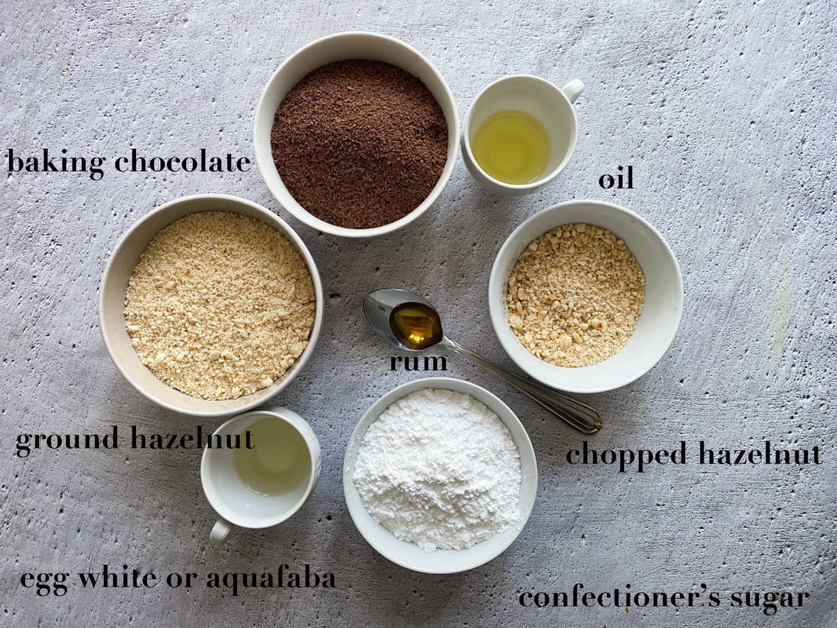 Ingredients on a white background: bowls filled with chocolate, hazelnuts, oil, eggwhite, sugar and spoon with rum.