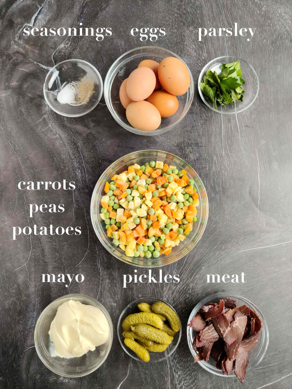 Seasonings, eggs, parsley, carrots, peas, potatoes, mayo, pickles and meat on a gray background. 