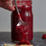 A slice of bread with jam, hand that's holding a spoon full of jam, and a jar in the background on gray background.
