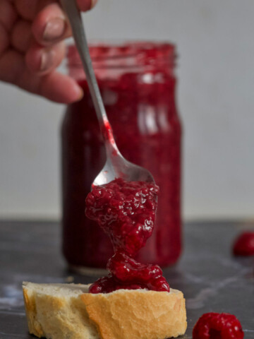 A slice of bread with jam, hand that's holding a spoon full of jam, and a jar in the background on gray background.