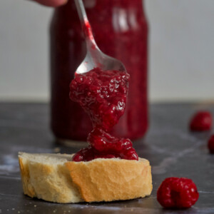 Spoon full of raspberry jam, a slice of bread and jar of raspberry jam.